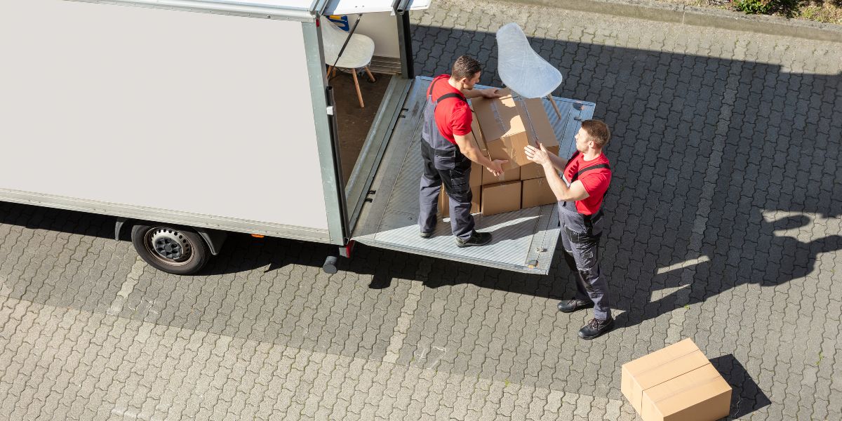 Two movers in uniform loading cardboard boxes onto a truck's lift gate.